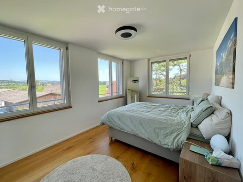 Bright bedroom with wooden flooring, large windows offering a scenic view, a neatly made bed, a nightstand with a lamp, and a round rug on the floor.