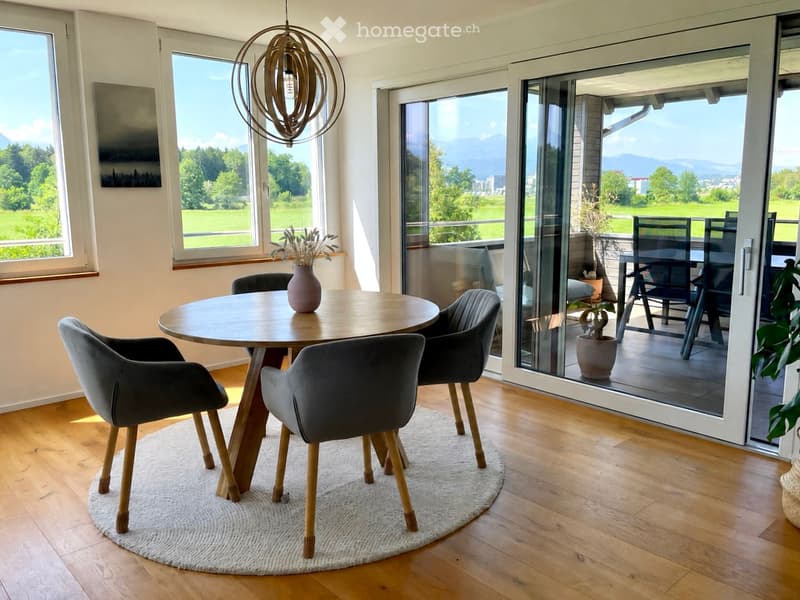 Modern dining area with a round wooden table, four grey chairs, a hanging light fixture, and a view of a green landscape through large windows and a sliding glass door leading to a balcony.