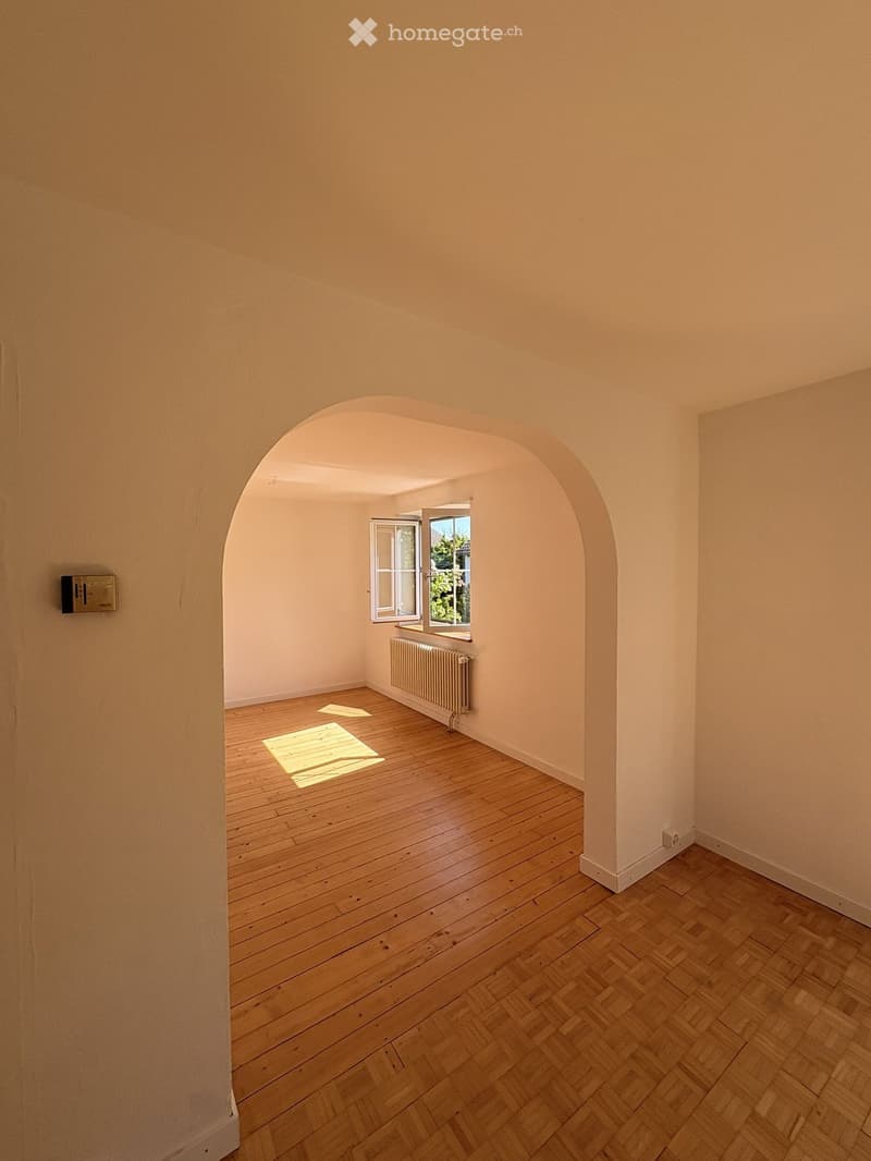 Empty room with wooden flooring, light-colored walls, and a radiator near a window with an arched doorway leading to another space.