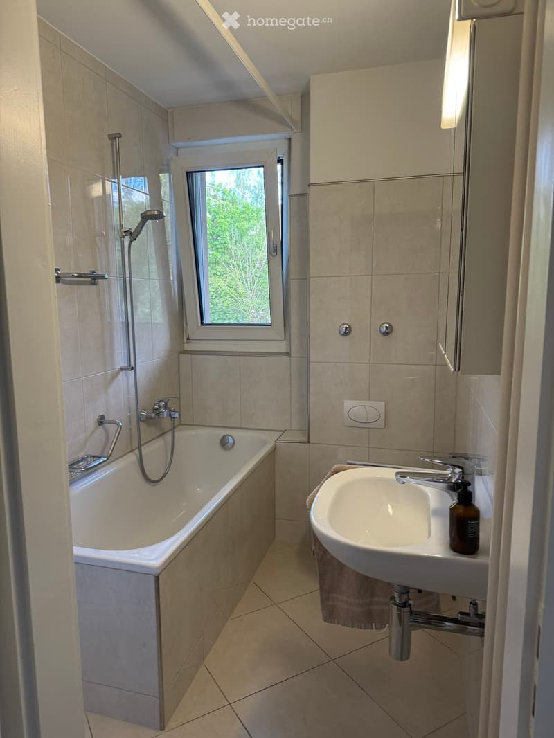 Modern bathroom with a bathtub, sink, showerhead, and a small window letting in natural light. Beige tiles and a towel are visible.