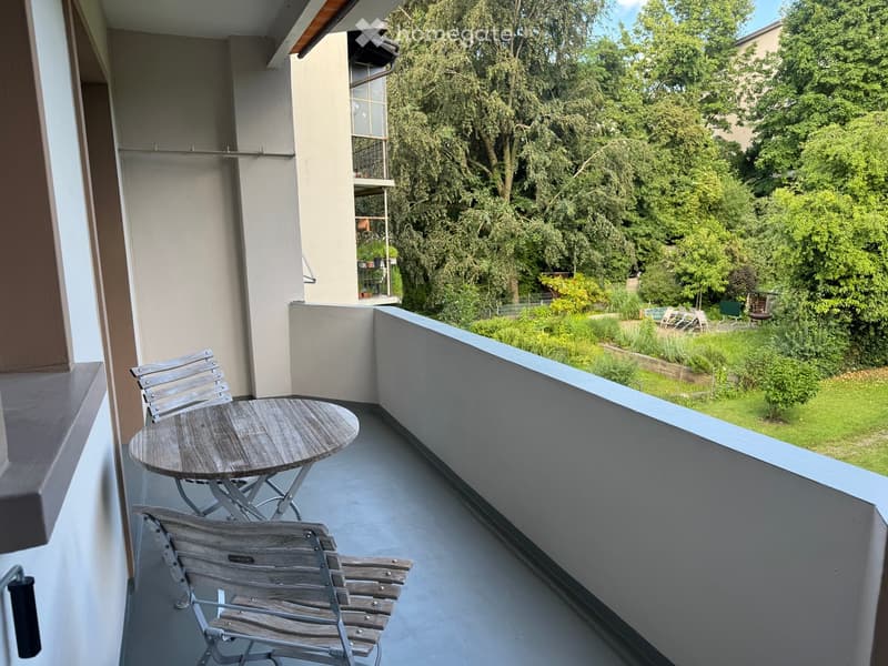 Balcony with a small round table and two wooden chairs, overlooking a green garden area with trees and lawn.