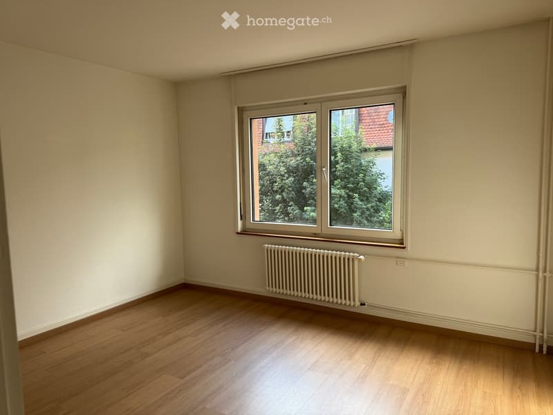 Empty room with wooden flooring, light-colored walls, a window with white frame, and a radiator below the window.
