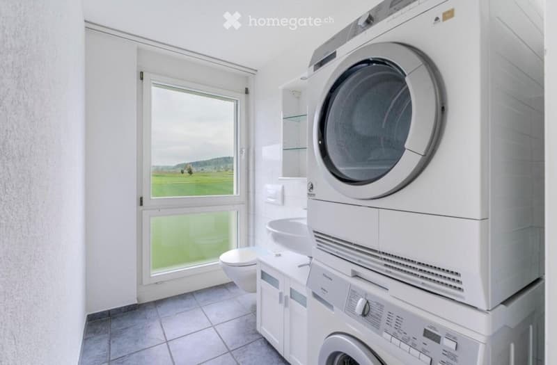 Bright bathroom with a toilet, sink, and stacked washing machine and dryer. Large window offers a scenic view of a green field.