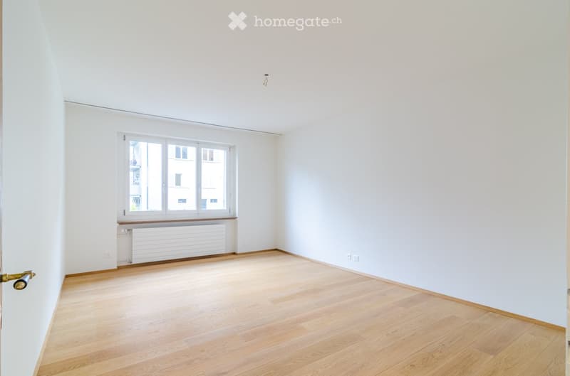 Empty room with light wooden flooring, white walls, and a large window with white frames. The room has a radiator below the window and a door with a brass handle on the left side.