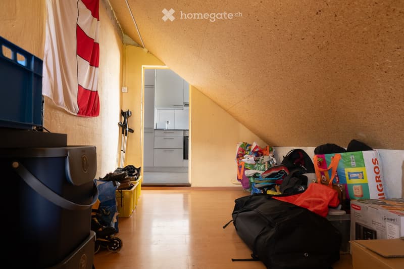 An attic room with wooden flooring, slanted ceilings, and exposed beams. Visible are kitchen cabinets, a sink, and drawers in the background, along with various stored items including suitcases, backpacks, shopping bags, and storage boxes.