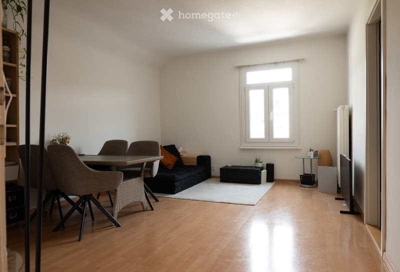A modern, well-lit living space with a sofa, dining table, chairs, and a plant. The room features light wooden flooring and white walls.