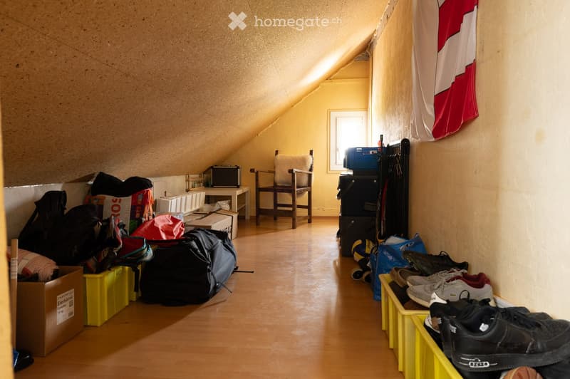 Attic room with wooden flooring, slanted ceilings, and various stored items including a chair, table, backpacks, suitcases, storage boxes, a radiator, a ball, and a shoe rack.