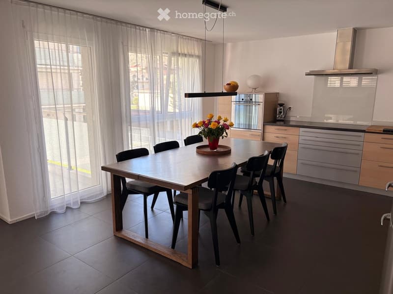 Modern kitchen with a large dining table, six chairs, a flower vase with yellow and pink flowers, a built-in oven, kitchen cabinets, a coffee machine, and a range hood.