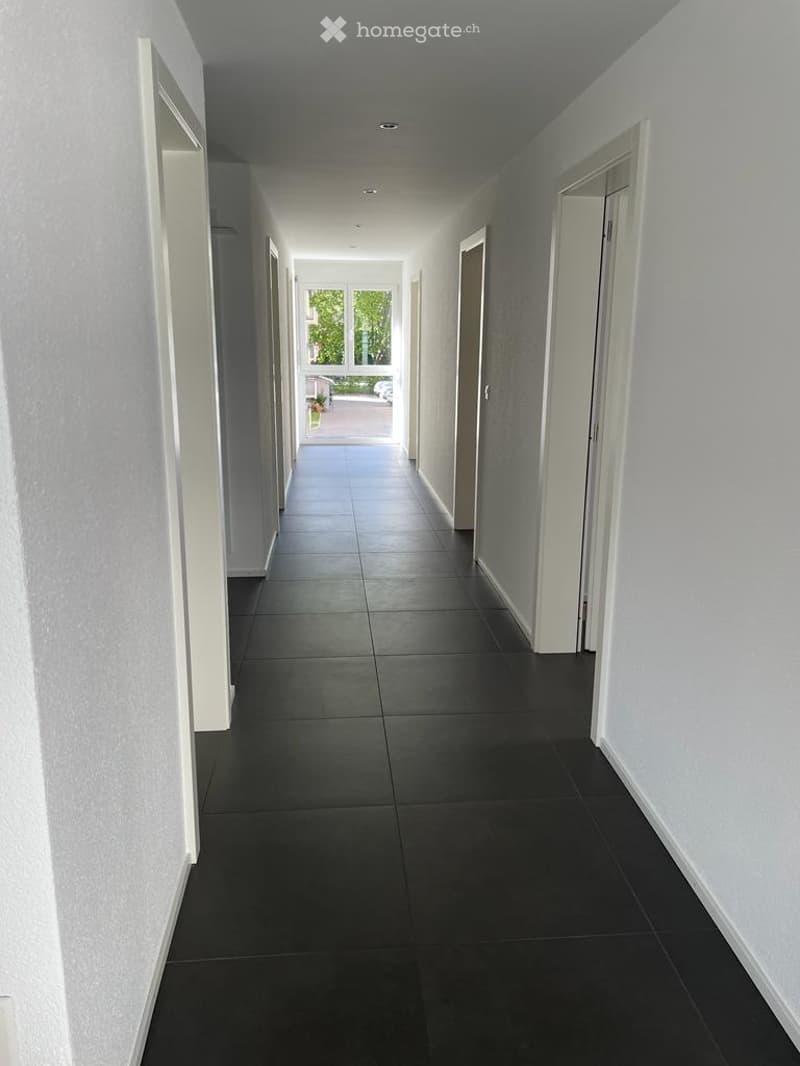 Clean, modern hallway with dark tiled flooring and white walls, leading to an open doorway with natural light.