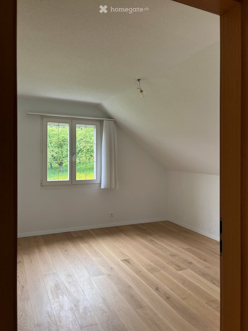 Empty attic room with wooden flooring, slanted ceilings, and a window with white curtains. The walls are painted in a light beige color.