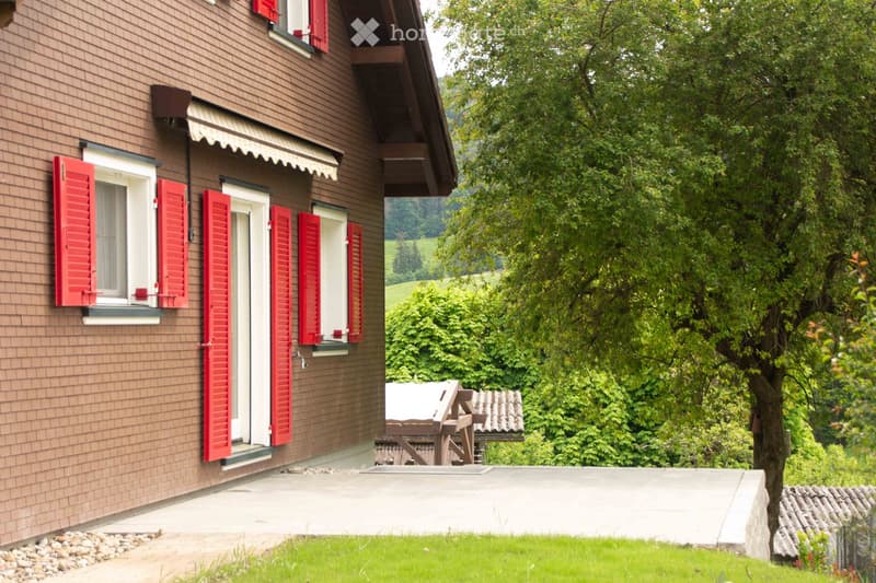 Two-story house with brown wooden siding, red shutters, and a white awning over the entrance. The house is surrounded by lush greenery and a well-maintained lawn.