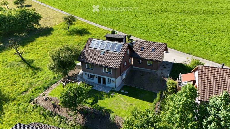 Aerial view of a two-story house with a brown roof and solar panels, surrounded by a well-maintained lawn and trees. The house features multiple windows and a small patio area.