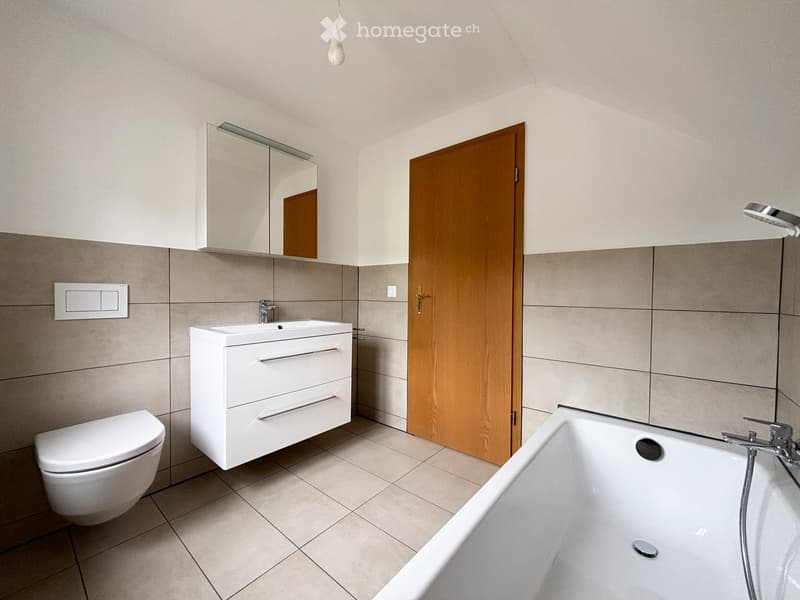 Modern bathroom with a white toilet, sink cabinet, bathtub, and a mirrored cabinet above the sink. The room features tiled walls and a wooden door.
