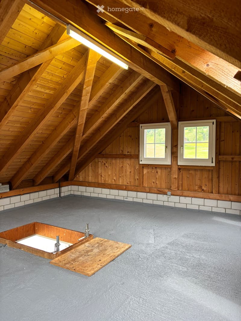 An attic space with wooden beams, exposed wooden ceiling, two windows, and a wooden floor with a small wooden platform and a built-in storage compartment.