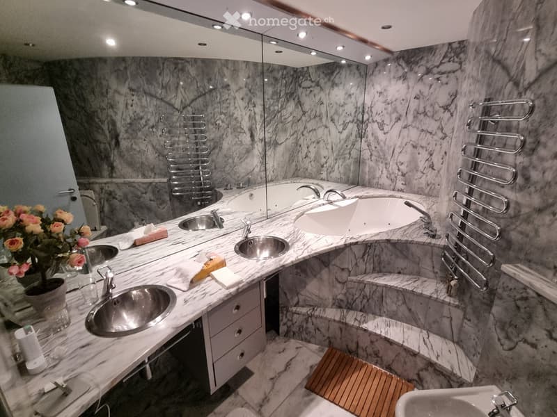 Luxurious bathroom with marble walls and floors, multiple sinks, a bathtub, towel racks, and a flower vase on the counter.