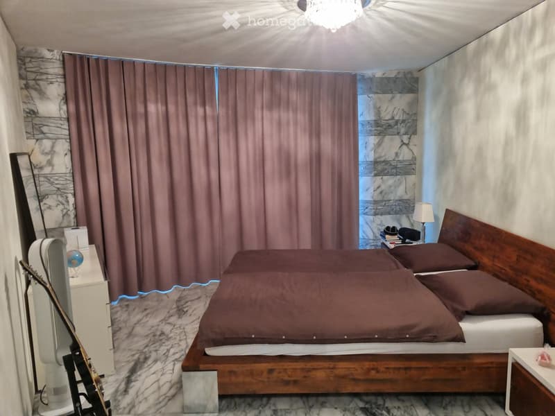 A modern bedroom with a large bed, wooden headboard, and brown bedding. Features include a guitar leaning against a white cabinet, a nightstand with a lamp, and large windows with brown curtains.