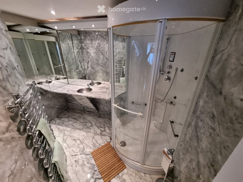 Luxurious bathroom with marble floors and walls, a large walk-in shower with glass enclosure, double sink vanity, and a towel rack with a green towel.