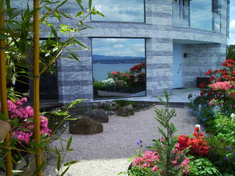 Modern house with large glass windows overlooking a scenic water view, surrounded by a well-maintained garden with colorful flowers and rocks.