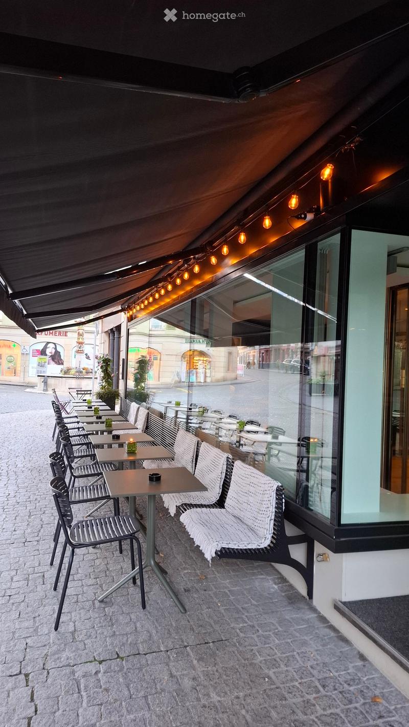 An outdoor seating area with tables and chairs, featuring a covered canopy with string lights, located on a cobblestone pavement next to a building with large windows.