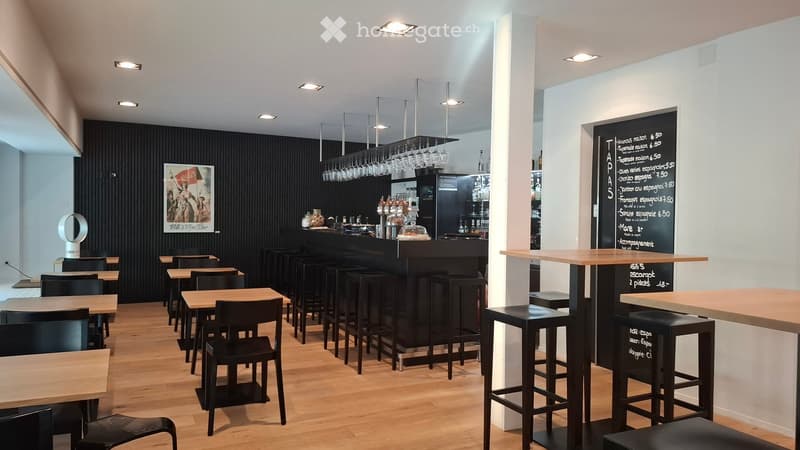 Modern bar area with wooden tables, black chairs, bar counter, hanging glasses, and a menu board.