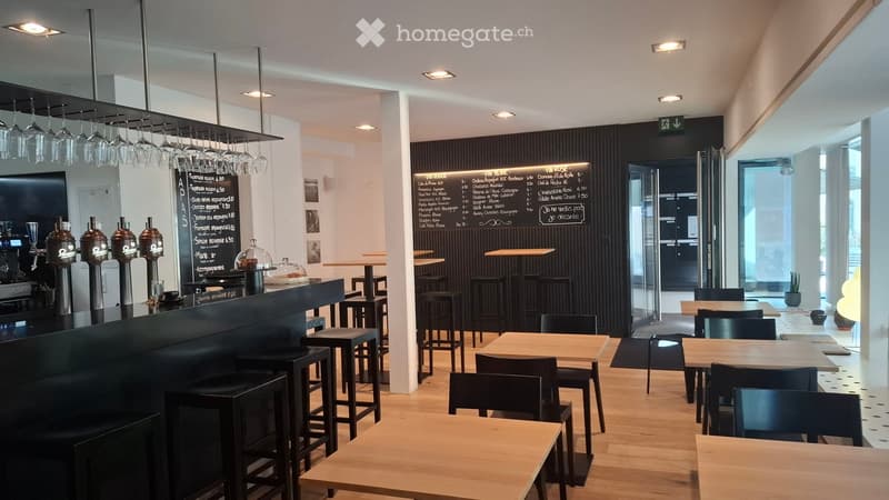 Modern restaurant interior with wooden tables, black chairs, a long bar counter, wine glasses hanging from the ceiling, and a menu board on the wall.