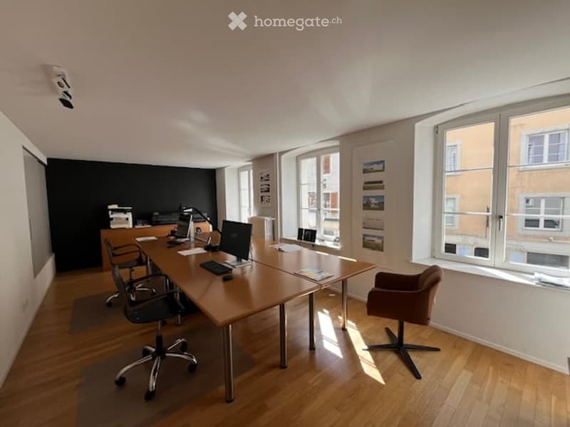 Bright, modern office space with wooden flooring, large windows, and a long wooden desk with office chairs, a laptop, keyboard, mouse, printer, and a lamp.