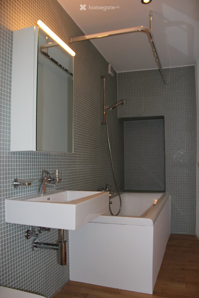 Modern bathroom with a white sink, chrome faucet, bathtub, and large mirror. Grey tiled walls and wooden flooring.