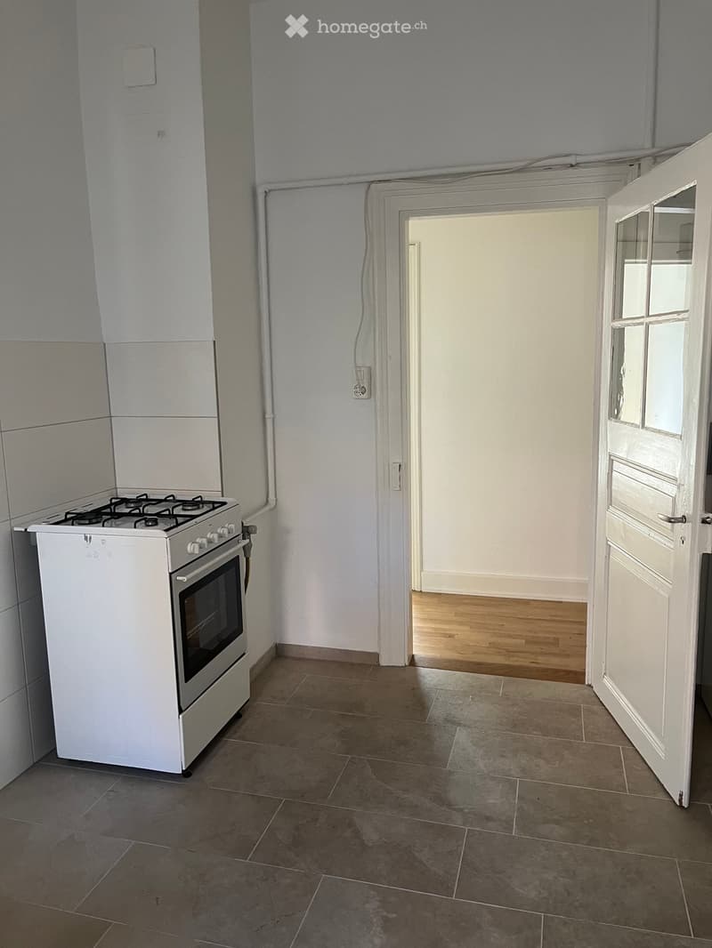 Empty kitchen with tiled floor, white stove, and open doorway leading to another room with wooden flooring.