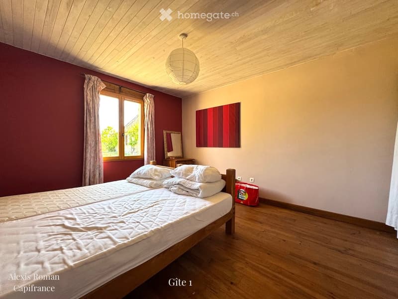 Cozy bedroom with wooden floor, large window with curtains, double bed with white bedding, mirror, and a decorative painting on the wall.