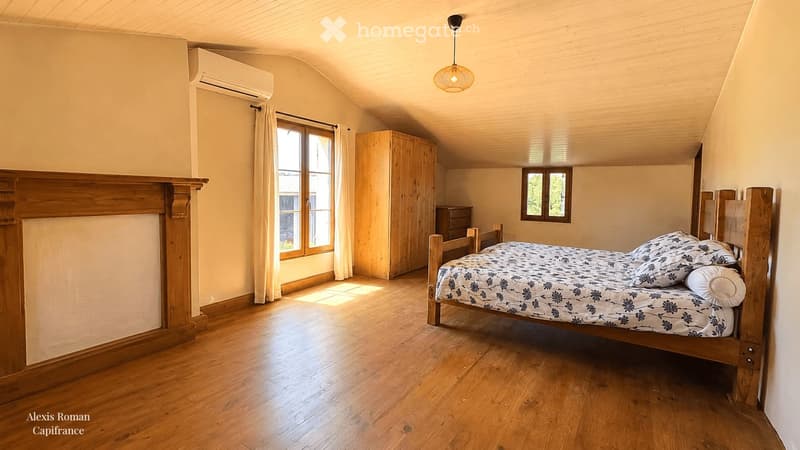 Cozy attic bedroom with wooden floors, slanted ceilings, and large windows. Features a wooden bed with patterned bedding, a wardrobe, a nightstand, and a pendant light.