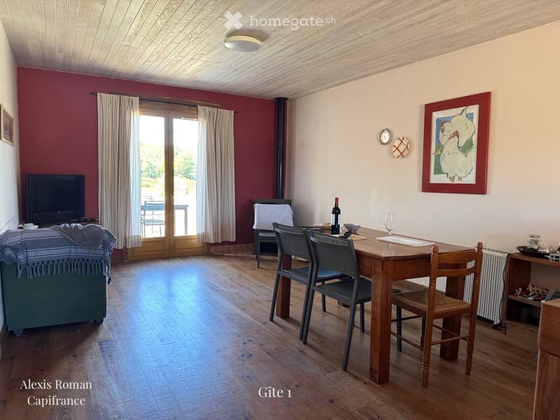 A cozy living and dining area with wooden flooring, a sofa, a dining table with chairs, a television, and a bottle on the table.