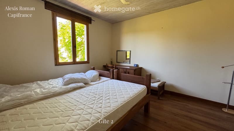Cozy bedroom with wooden bed, mirror, dresser, and chair. Large window with wooden frame lets in natural light.
