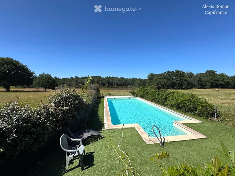 Outdoor area with a swimming pool, surrounded by greenery, featuring a lounge chair and a plastic chair on artificial grass.