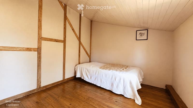 Minimalist bedroom with wooden floor, white walls, a single bed with white linens, and a small woven throw on top. A simple picture frame hangs on the wall.