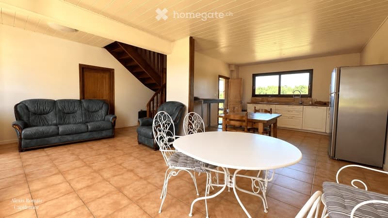 Open-plan living and dining area with a sofa, armchair, dining table, chairs, and a kitchen area with a refrigerator and counter.