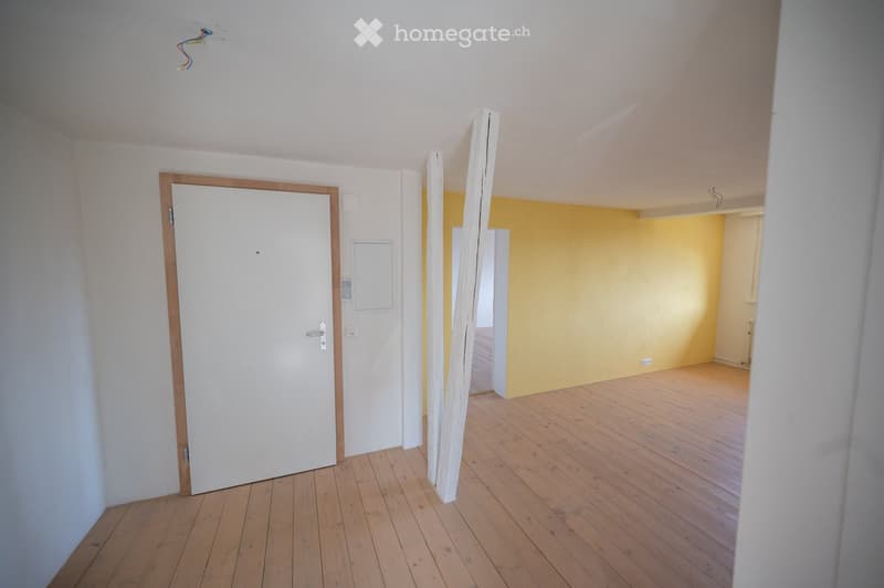 Empty room with wooden flooring, white walls, and a white door with a wooden frame. The adjacent room has yellow walls and wooden flooring.