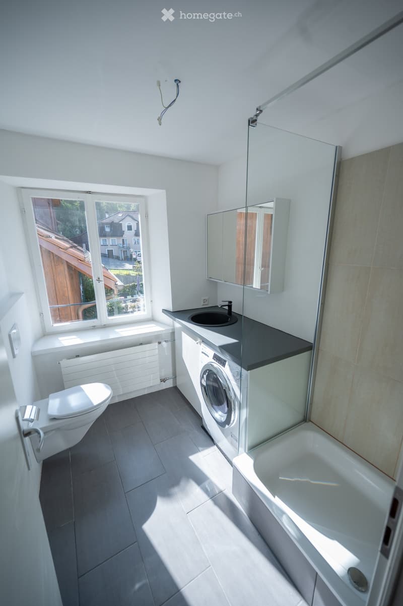 Modern bathroom with a toilet, sink, bathtub, washing machine, and a large window offering a view of the garden.