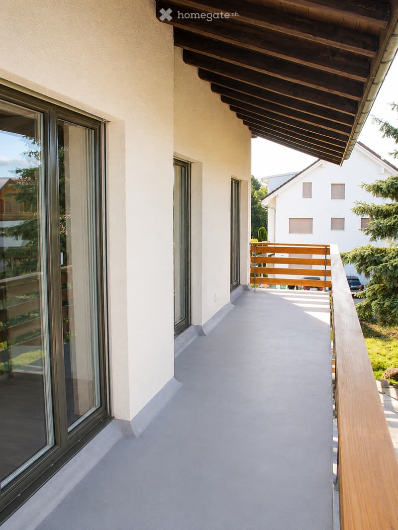 Balcony with wooden railing, large windows, and a covered wooden roof extending over the balcony.