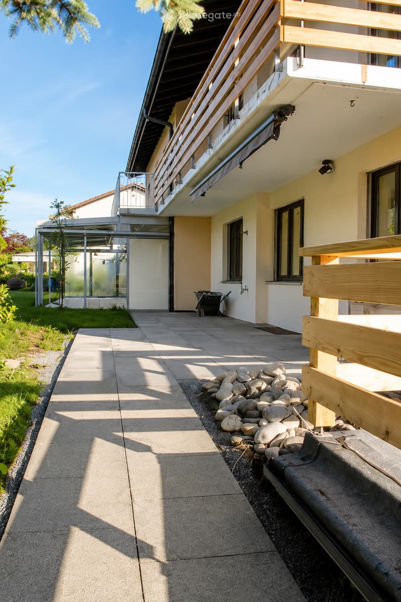 A modern outdoor area with a paved walkway, wooden balcony railing, and decorative rocks. The building has a light yellow facade with large windows and a covered entrance area.