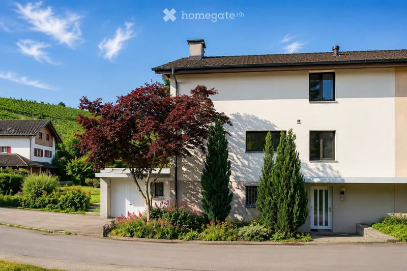 Two-story house with white exterior, multiple windows, and a chimney. The house is surrounded by well-maintained landscaping, including trees and shrubs, with a driveway leading to the entrance.