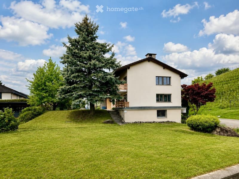 Two-story house with a raised ground floor, surrounded by a well-maintained lawn and trees, including a prominent conifer and a red-leaved tree. The house features a balcony and multiple windows.