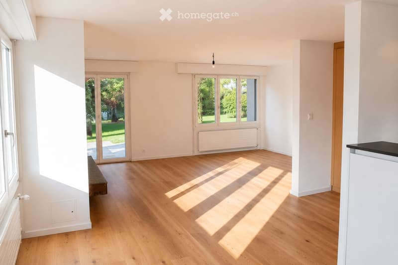 Empty room with light wooden flooring, white walls, large windows, and a sliding glass door leading to a garden.