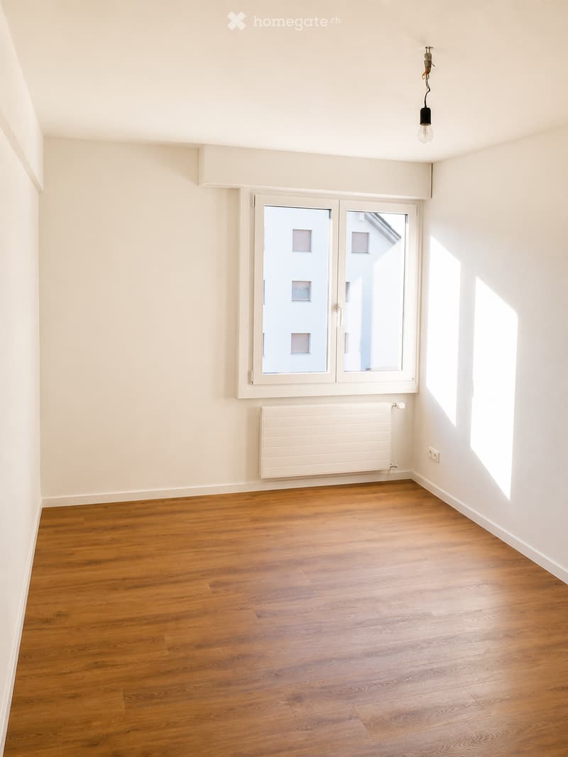 Empty room with wooden flooring, white walls, a window with white frame, a radiator, and a single light fixture on the ceiling.