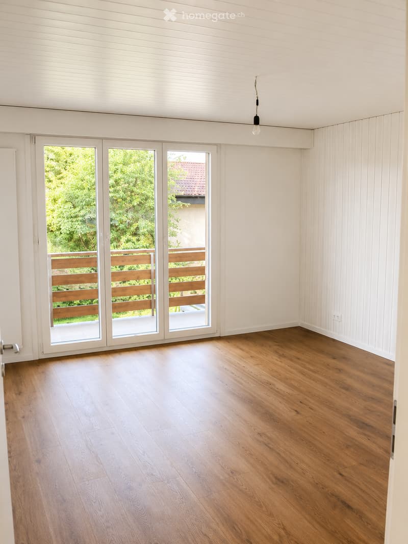 Empty room with wooden flooring, white walls, large sliding glass doors leading to a balcony, and a hanging light fixture.