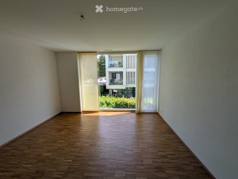 Empty room with wooden flooring, large window with blinds, and visible view of a neighboring balcony and greenery.