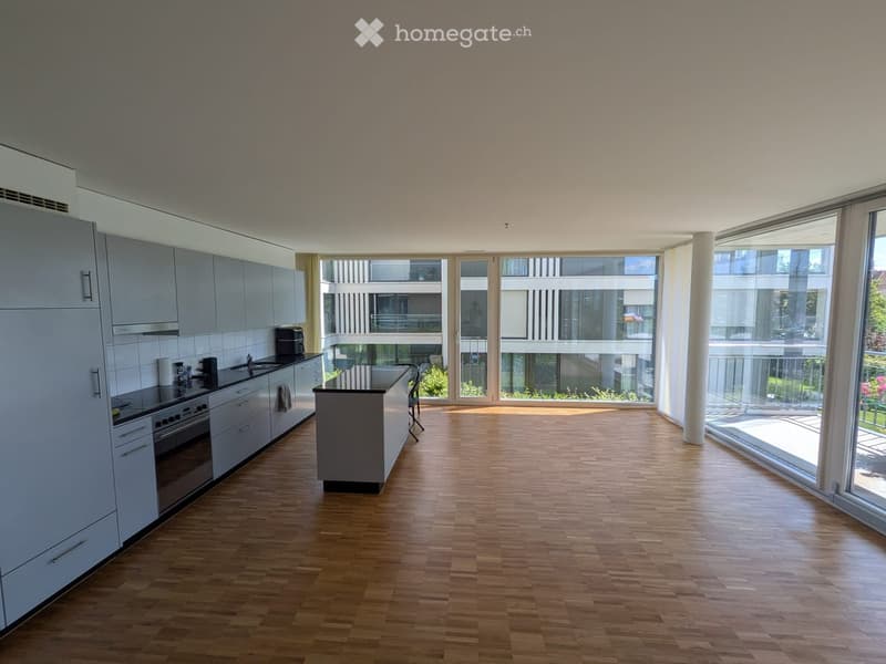 Modern kitchen with light grey cabinets, wooden flooring, and large windows overlooking a balcony area.