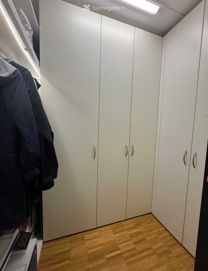 A narrow hallway with white built-in wardrobes and wooden flooring. A few jackets are hanging on the left side.