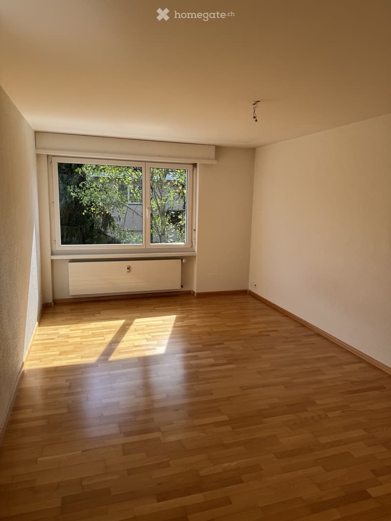 Empty room with wooden flooring, white walls, large window with blinds, radiator, and visible electrical outlets.
