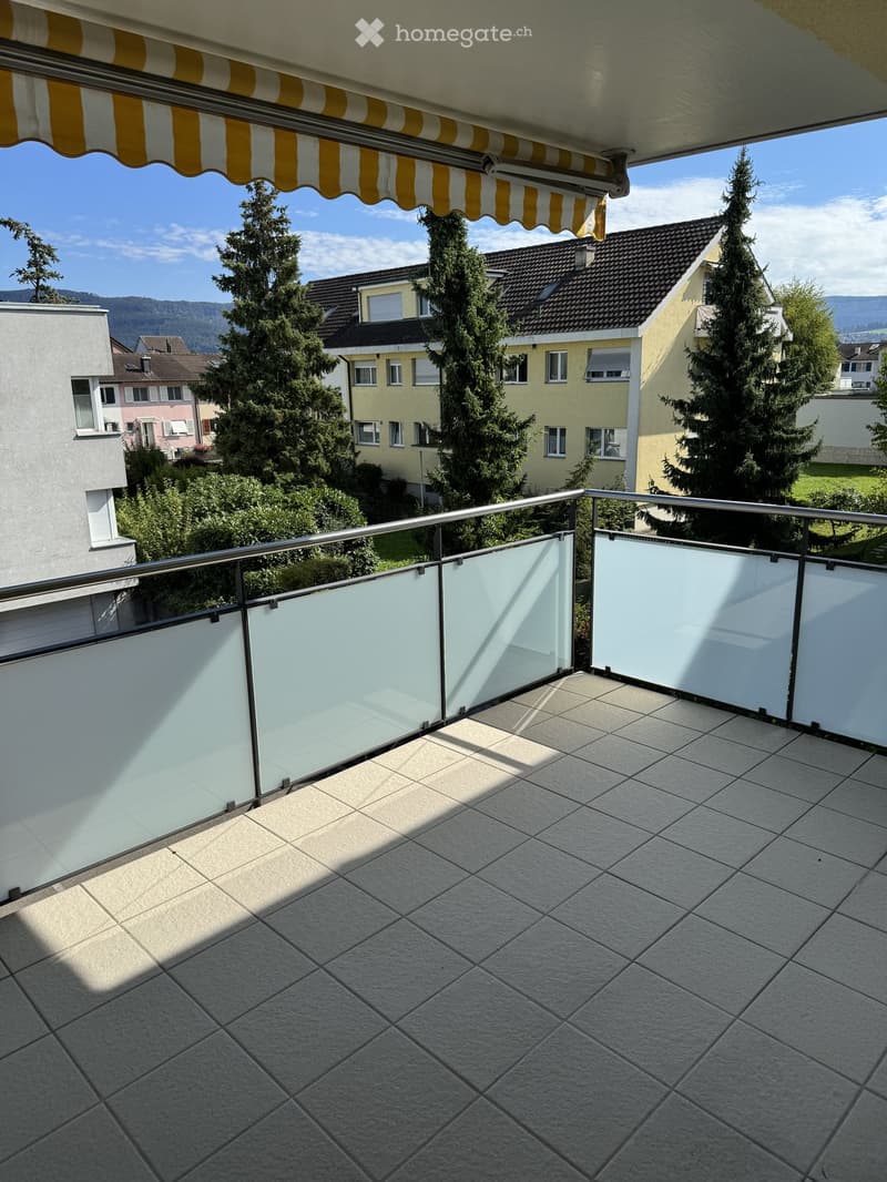 Balcony with tiled floor, glass railing, and a retractable awning, overlooking a residential area with trees and houses.