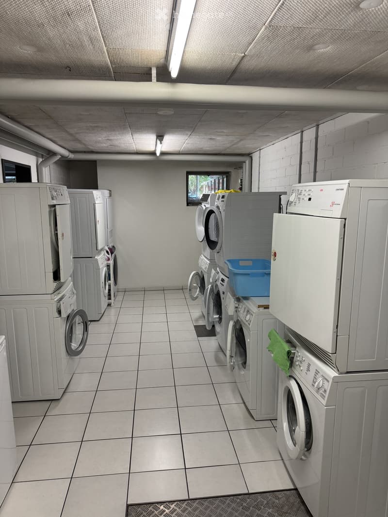 Laundry room with multiple washing machines and dryers, tiled floor, and a small window showing greenery outside.
